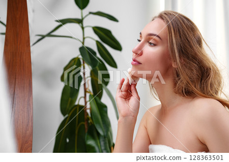 Woman applying facial treatment while looking in mirror in modern bathroom during daytime 128363501