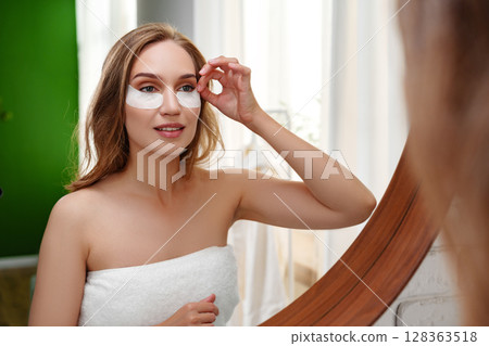 Woman applying under-eye patches in a bright, relaxing bathroom setting during daytime Woman applying under-eye patches in a bright, relaxing bathroom setting during daytime 128363518