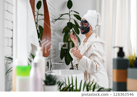 Woman applies charcoal face mask in modern bathroom during self-care routine in the afternoon 128363546