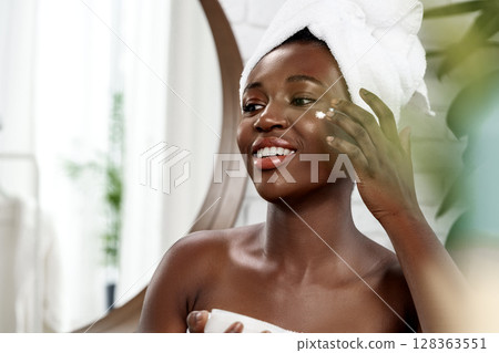 Woman applying moisturizer in a bright bathroom with a towel on her head during skincare routine 128363551