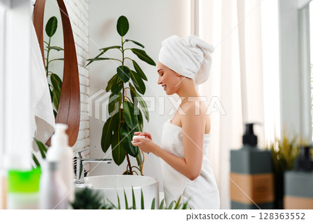 Woman in towel applying skincare product in modern bathroom near window with plants 128363552