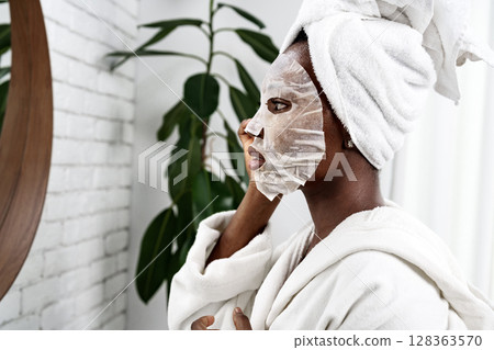 Woman applying a face mask while looking in the mirror at home spa setting during morning routine 128363570