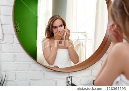 Woman applying skincare in bathrobe while looking in mirror in a bright bathroom during morning routine Woman applying skincare in bathrobe while looking in mirror in a bright bathroom during morning routine 128363593