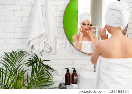 Woman applies skincare in a bright bathroom with plants and a mirror during morning routine 128363594