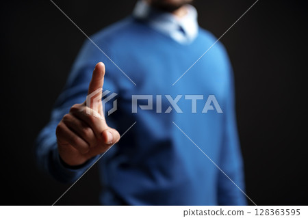 Man signaling to pause with one finger raised, dressed in casual attire against a dark background with soft lighting 128363595