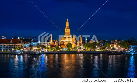 Night view of Wat Arun temple beautifully illuminated on the Chao Phraya River with glowing reflections and city lights in the background 128363615