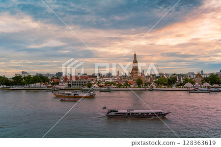 Evening view of Wat Arun temple across the Chao Phraya River with boats passing by under a dramatic sunset sky 128363619