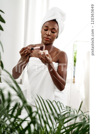 Woman applying makeup in a bright bathroom featuring plants and natural light during the day 128363649