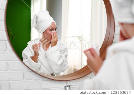 Woman applying skincare in bathrobe while looking in mirror in a bright bathroom during morning routine Woman applying skincare in bathrobe while looking in mirror in a bright bathroom during morning routine 128363654