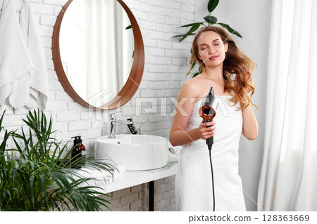 Woman drying her hair with a hair dryer in a modern bathroom with plants, mirror, and white decor Woman drying her hair with a hair dryer in a modern bathroom with plants, mirror, and white decor 128363669