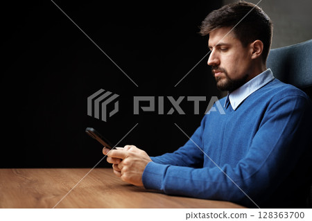 Man in blue sweater sitting at a desk focused on his smartphone during evening hours 128363700