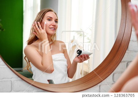 Woman applying skincare product in bathroom mirror during morning routine with natural light enter 128363705