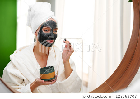 Woman applies charcoal face mask in spa-like setting during self-care routine at home in natural light 128363708