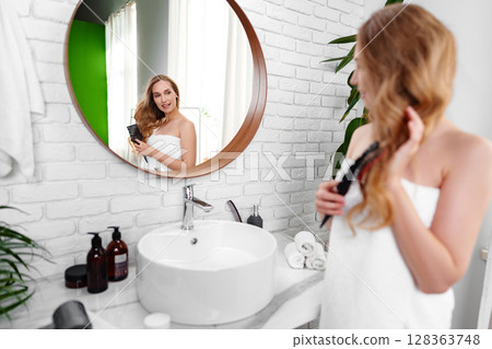 Woman grooming hair in a modern bathroom with natural light and stylish decor 128363748