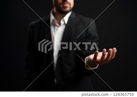 Businessman in formal attire extends hand in welcoming gesture against dark background during networking event Businessman in formal attire extends hand in welcoming gesture against dark background during networking event 128363750