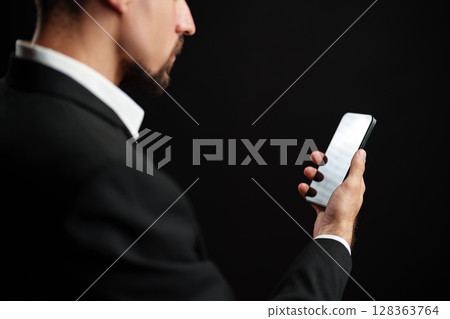 Man in formal attire checking smartphone against a dark background in a modern setting 128363764