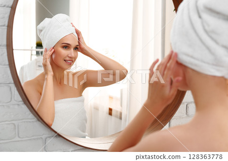 Woman in a towel preparing for the day while admiring herself in a mirror in a bright bathroom at morning time Woman in a towel preparing for the day while admiring herself in a mirror in a bright bathroom at morning time 128363778