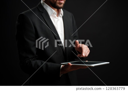 Professional man in suit using tablet while standing against dark background indoors 128363779