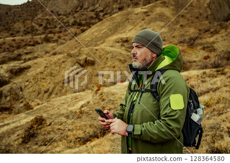 Man standing on rocky terrain in rugged outdoor gear while checking his phone during an adventure in nature Man standing on rocky terrain in rugged outdoor gear while checking his phone during an adventure in nature 128364580