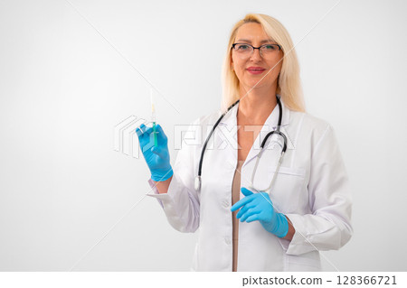 Blonde woman doctor wearing white coat, gloves, and stethoscope holding a single syringe. Clean studio shot on white background with copy space 128366721