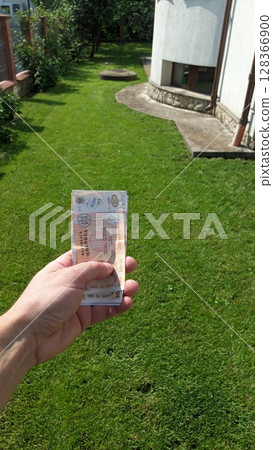 A Hand Holding Currency in a Beautiful Garden Setting Surrounded by Lush Greenery and Nature 128366900