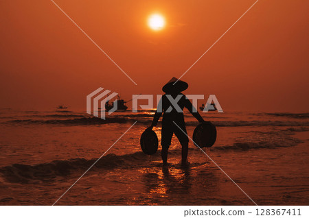 Silhouette of a Vietnamese woman in a straw hat on shore of sea with ships in a fishing village in the morning at sunrise in Vietnam Silhouette of a Vietnamese woman in a straw hat on shore of sea with ships in a fishing village in the morning at sunrise in Vietnam 128367411