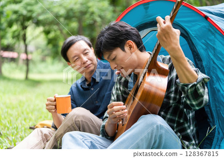 Family enjoyment during camping. Father watching son play guitar in a tent. Family enjoyment during camping. Father watching son play guitar in a tent. 128367815