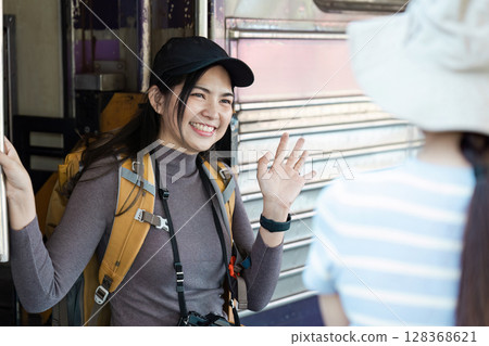 Young Female Traveler with Backpack and Camera Greeting Friend at Train Station, Ready for Adventure 128368621