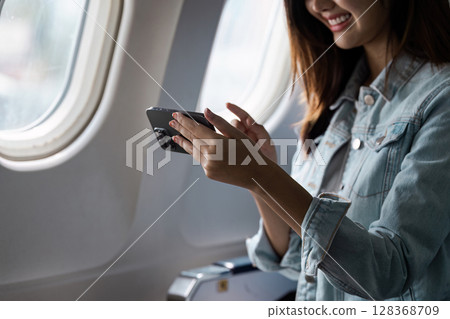 Young Woman Using Smartphone by Airplane Window During Flight, Enjoying Travel Experience Young Woman Using Smartphone by Airplane Window During Flight, Enjoying Travel Experience 128368709