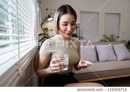Young Woman Taking Medication with Water in a Modern Living Room Setting, Promoting Health and Wellness 128368710