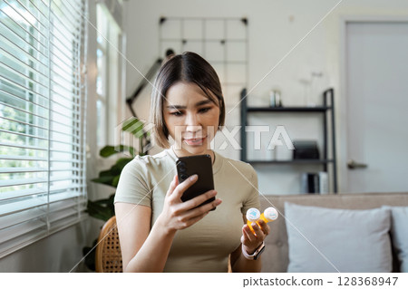 Young Woman Using Smartphone for Online Healthcare Consultation While Holding Medication in Modern Home Setting Young Woman Using Smartphone for Online Healthcare Consultation While Holding Medication in Modern Home Setting 128368947