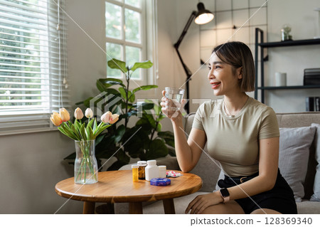 Young Woman Taking Medication at Home, Sitting on Sofa with Glass of Water, Surrounded by Healthcare Products and Natural Light Young Woman Taking Medication at Home, Sitting on Sofa with Glass of Water, Surrounded by Healthcare Products and Natural Light 128369340