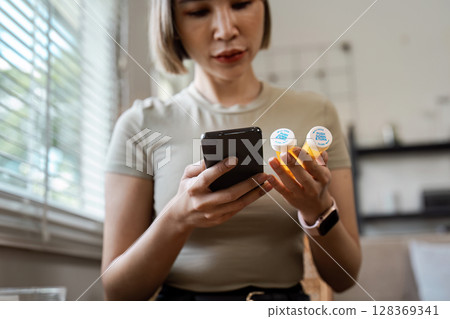 Woman Checking Medication Information on Smartphone While Holding Pill Bottles in a Modern Home Setting Woman Checking Medication Information on Smartphone While Holding Pill Bottles in a Modern Home Setting 128369341