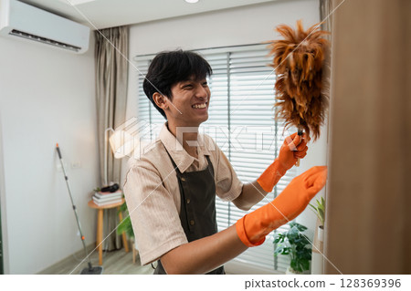 Young Man Smiling While Dusting Furniture in Modern Living Room with Orange Gloves and Feather Duster Young Man Smiling While Dusting Furniture in Modern Living Room with Orange Gloves and Feather Duster 128369396