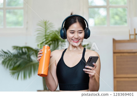 Young Woman Enjoying Wellness at Home with Headphones, Water Bottle, and Smartphone in a Modern Living Room 128369401