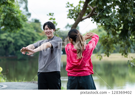 Young Couple Stretching Before Jogging in a Scenic Park with Lush Greenery and a Serene Lake in the Background 128369402