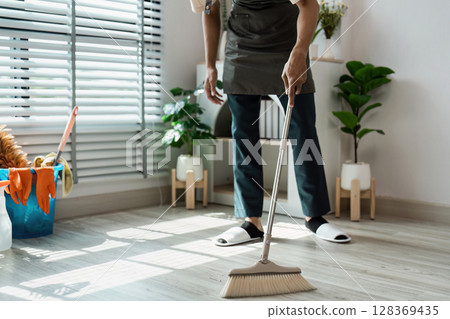 Man Sweeping Floor in Modern Living Room with Plants and Sunlight Through Blinds Man Sweeping Floor in Modern Living Room with Plants and Sunlight Through Blinds 128369435