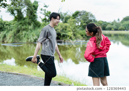 Young Couple Stretching and Chatting Before Jogging by a Scenic Lake in a Park 128369443