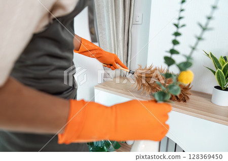 Close-up of Person Cleaning with Feather Duster and Orange Gloves in Modern Home Interior 128369450