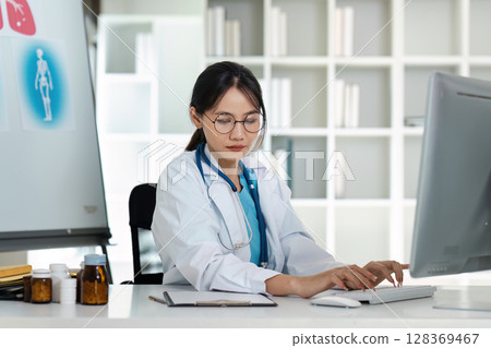 Female Doctor Working on Computer in Modern Medical Office with Medical Equipment and Bookshelves in Background Female Doctor Working on Computer in Modern Medical Office with Medical Equipment and Bookshelves in Background 128369467