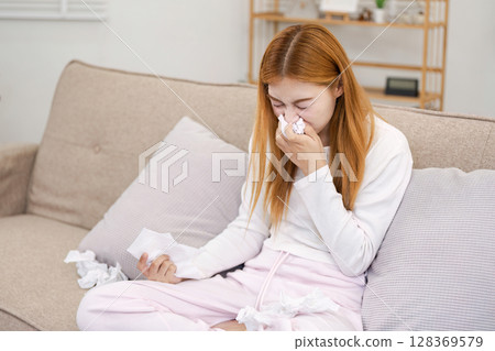 Young Woman Feeling Sick at Home, Sitting on Couch with Tissues, Blowing Nose and Looking Unwell 128369579