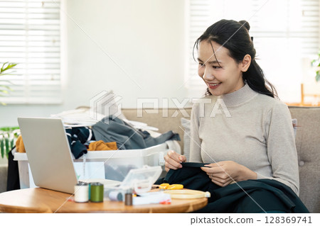 Woman Recycling Clothes at Home with Sewing Kit and Laptop, Promoting Sustainable Fashion and Eco-Friendly Practices Woman Recycling Clothes at Home with Sewing Kit and Laptop, Promoting Sustainable Fashion and Eco-Friendly Practices 128369741
