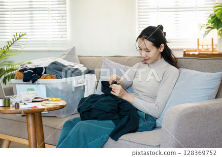 Woman Recycling Clothes at Home by Sewing and Mending on a Comfortable Sofa in a Bright Living Room Woman Recycling Clothes at Home by Sewing and Mending on a Comfortable Sofa in a Bright Living Room 128369752