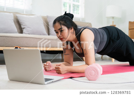 Overweight Woman Engaging in Home Workout for Wellness and Fitness with Laptop and Dumbbell Overweight Woman Engaging in Home Workout for Wellness and Fitness with Laptop and Dumbbell 128369781
