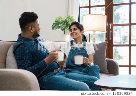 Happy Indian Couple Relaxing at Home, Enjoying Coffee on a Cozy Sofa in a Modern Living Room 128369824