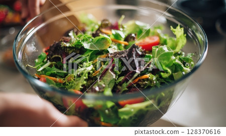 First-person view of assembling a fresh salad in a glass bowl with vibrant greens and vegetables 128370616