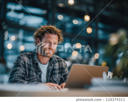 Man working on laptop in modern office with blurred background and warm lighting creating focused atmosphere 128374311