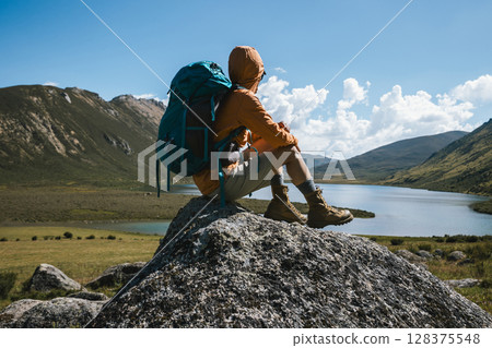 Backpacking woman sit on high altitude mountain top,enjoy the view of a lake in the distance Backpacking woman sit on high altitude mountain top,enjoy the view of a lake in the distance 128375548