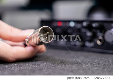 Human hand holds an XLR audio cable connector, poised for connection to a blurred professional sound interface, showcasing audio engineering and recording Human hand holds an XLR audio cable connector, poised for connection to a blurred professional sound interface, showcasing audio engineering and recording 128377887