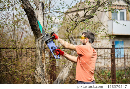 Young man cutting trees using electrical chainsaw in motion at his backyard 128379247